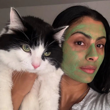Acne care routine with a woman wearing a green face mask holding a black and white cat indoors